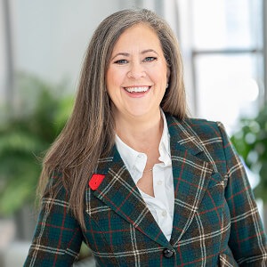 A woman with long light brown hair smiles and wears a plaid green jacket and a white shirt. 