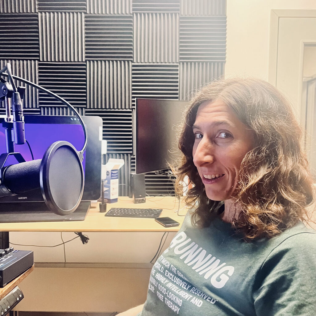 A white woman with wavy brown hair and a gray t-shirt, smiling slightly, sits a recording studio
