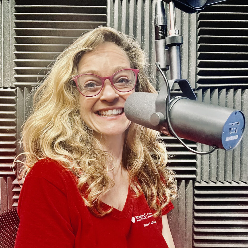 A white woman behind a microphone has blonde hair, red glasses, and wears a bright red t-shirt.