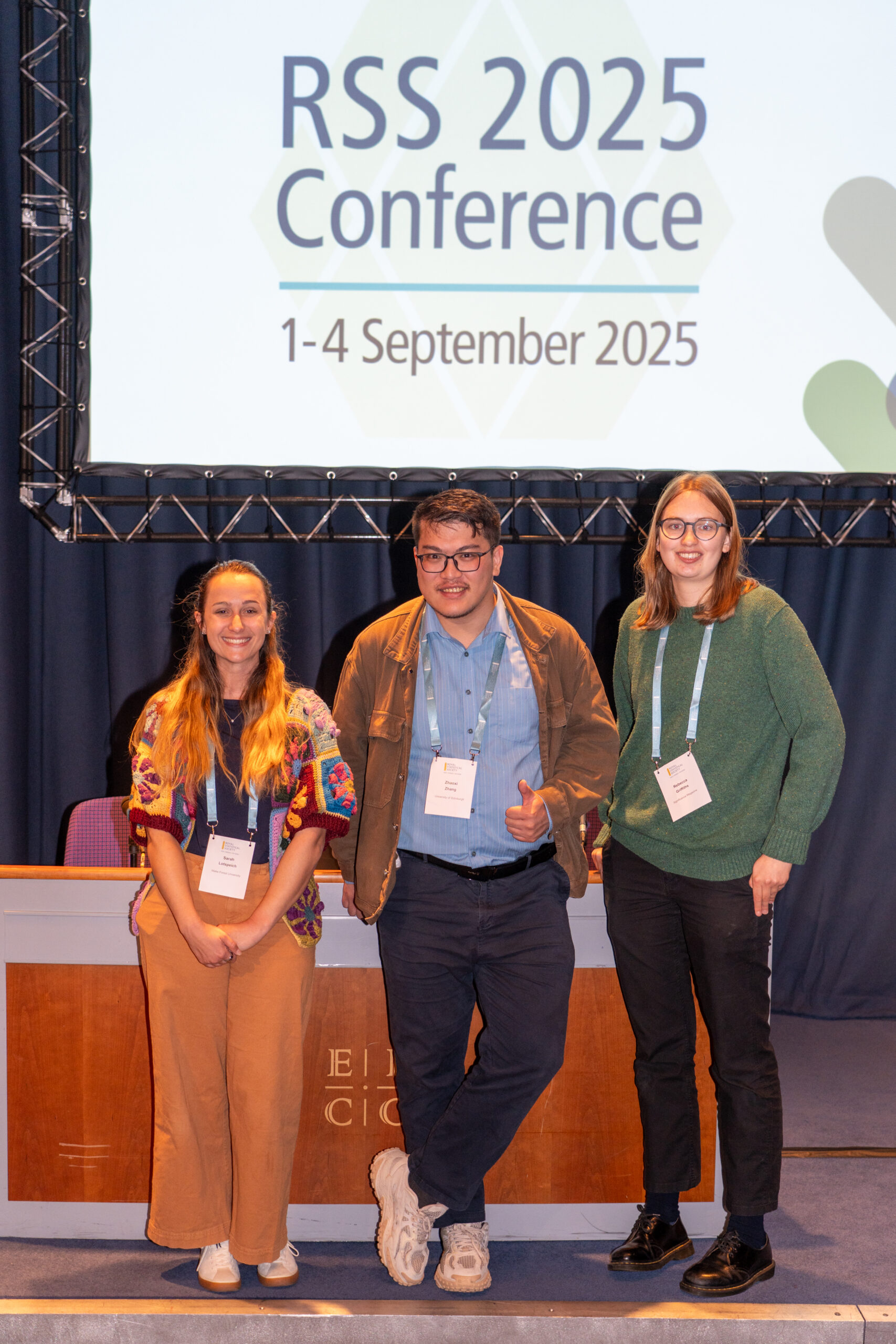 Two women and one man, in the middle, in business casual, with nametags, stand under a sign reading RSS 2025 Conference 1-4 September 2025