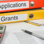 Binders labeled "applications" and "Grants" with writing tools in the foreground