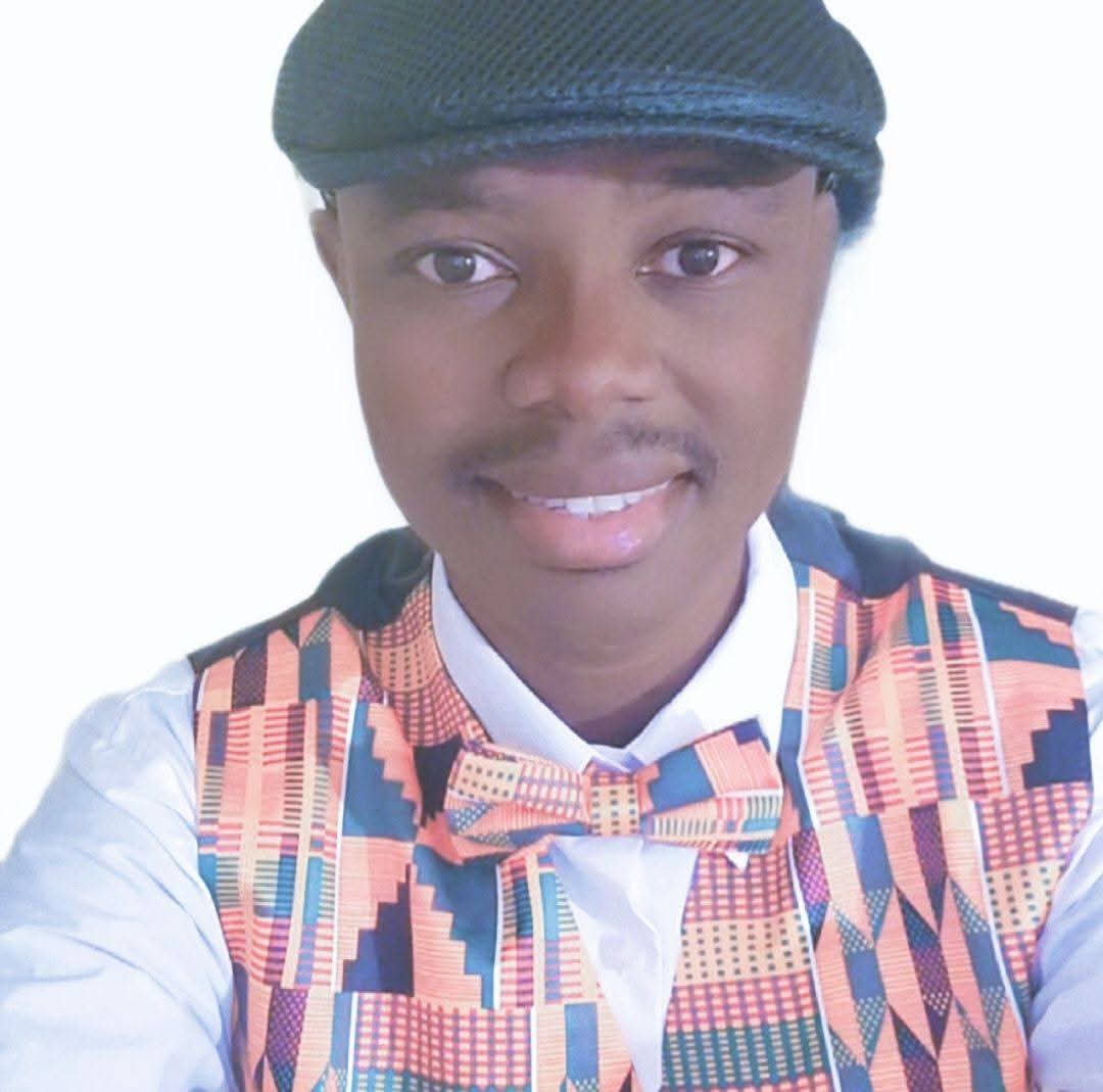 Timothy Boakye smiles at the camera in a black page boy hat, a kente-patterned vest and bowtie, and a white shirt.