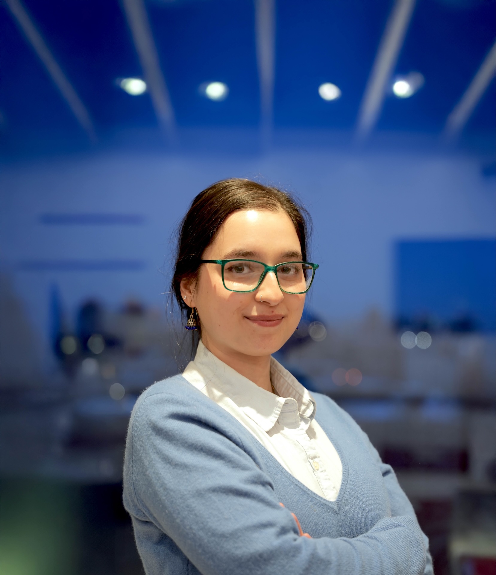 Adeeba Tak has brown hair, glasses, and is wearing a light blue sweater with a white shirt.