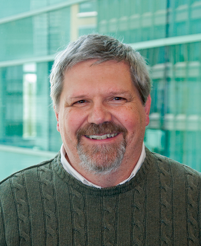 David Corliss has short gray hair, a short beard, and is wearing a green cable-knit sweater.