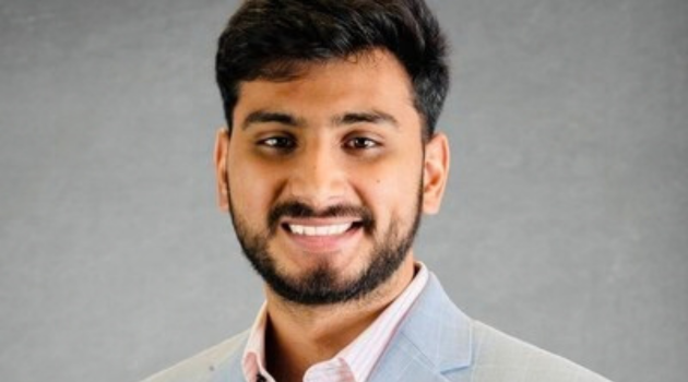 Karan Kumar is wearing a pale blue suit jacket with a light pink shirt. He has a short beard and short, dark hair.